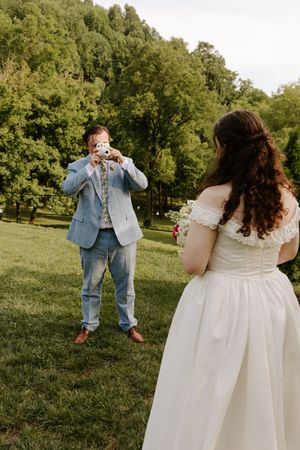 Groom in light blue suit photographs bride in white off-shoulder wedding dress outdoors on green lawn.