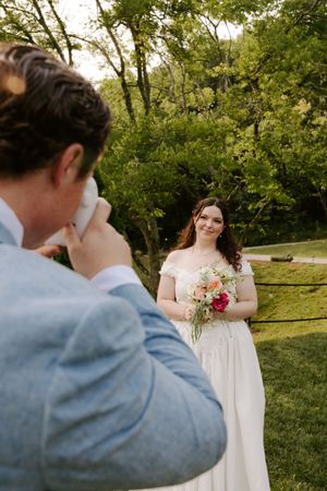 Bride in white gown smiles holding pink bouquet while groom photographs her in lush garden setting.