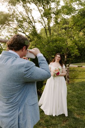 Groom in blue suit photographing bride in white gown holding colorful bouquet outdoors among lush green trees.