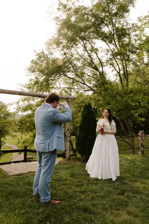 Groom photographs smiling bride holding bouquet near rustic wooden arch in lush garden setting.