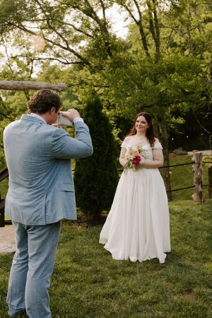 Bride in white off-shoulder gown holds bouquet while groom in light blue suit photographs her outdoors.