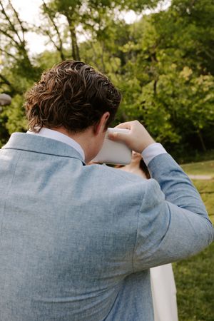 Groom in light blue suit photographing with instant camera outdoors at wedding reception.