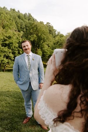 A bride photographs her groom in a light blue suit with a floral boutonniere on a green lawn at their outdoor wedding.