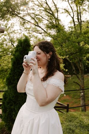 Bride in white off-shoulder dress photographs outdoors with Instax camera, surrounded by lush green trees.