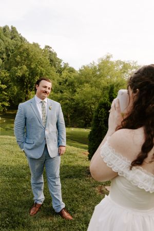Bride photographs groom in light blue suit and floral tie on green lawn at outdoor wedding.
