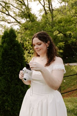 Bride in white off-shoulder dress holding Instax camera in lush garden setting.
