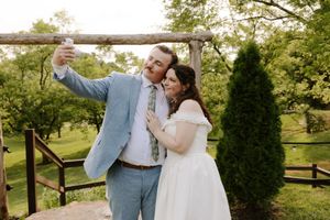 Bride and groom taking a selfie outdoors near a rustic wooden arbor surrounded by lush green trees.