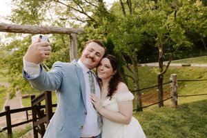 Bride and groom taking a selfie outdoors near rustic wooden fence surrounded by lush green trees on wedding day.