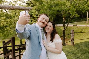 Bride and groom taking a wedding selfie outdoors, smiling together near a wooden fence surrounded by green trees.