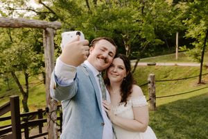 Bride and groom take a joyful selfie outdoors at their wedding venue surrounded by lush greenery.