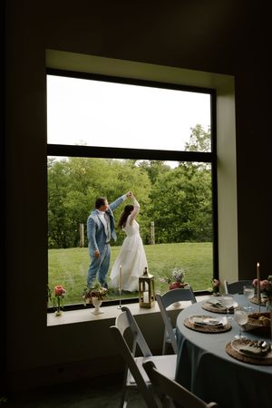Bride and groom dancing outdoors viewed through large window with reception table.