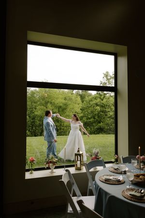 Bride and groom dance on green lawn viewed through large window with elegant reception table inside.