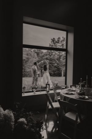 Black and white photo of a bride and groom dancing outside a window, viewed from a decorated reception venue.