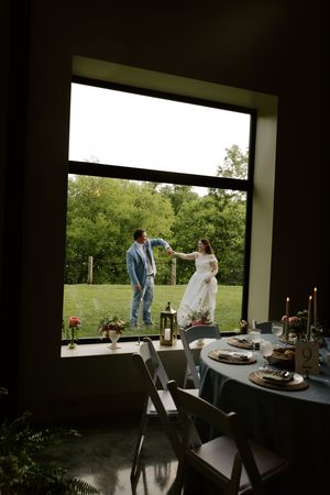 Bride and groom share a romantic moment outside a large window, viewed from a candlelit reception table inside.