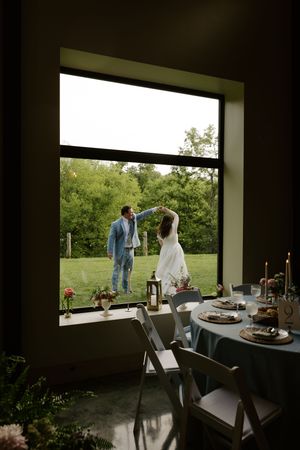 Bride and groom dancing outdoors viewed through large window with elegant reception table in foreground.