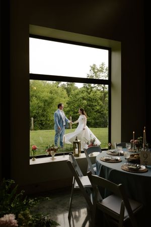 Bride and groom holding hands on green lawn viewed through large window beside elegant wedding reception table.