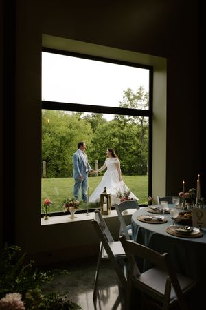 Wedding couple holds hands outside viewed through large window with reception table in foreground.