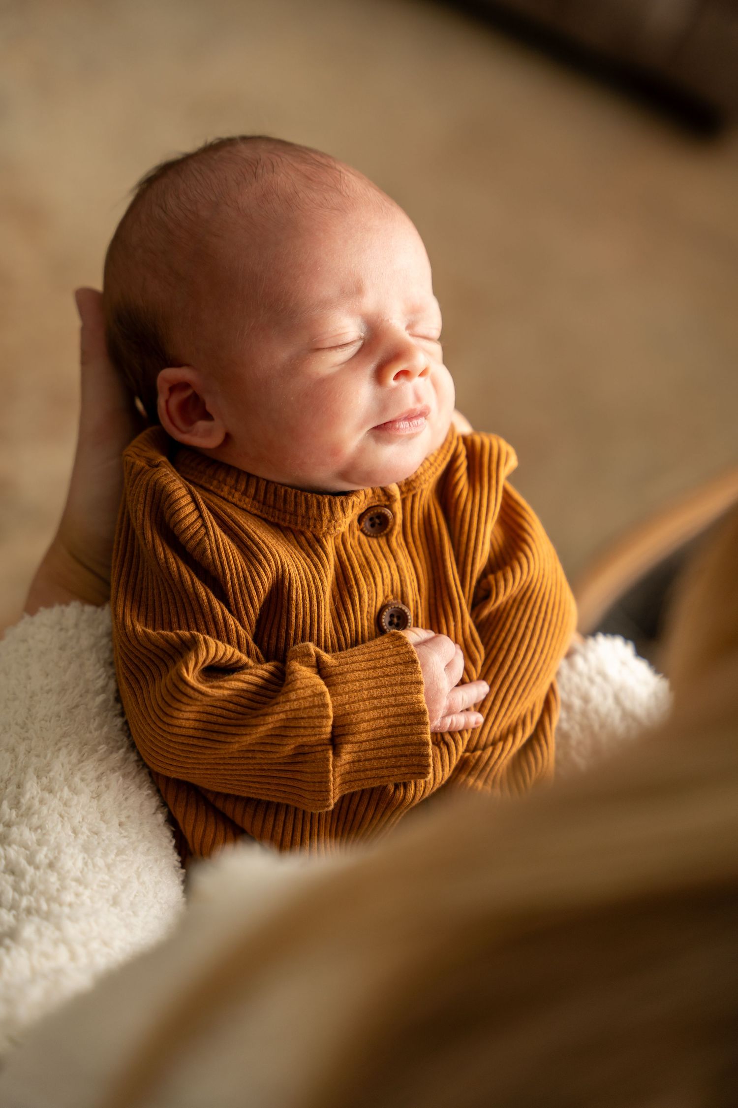 Sleeping newborn baby in a mustard yellow ribbed onesie resting peacefully on a white fluffy blanket.