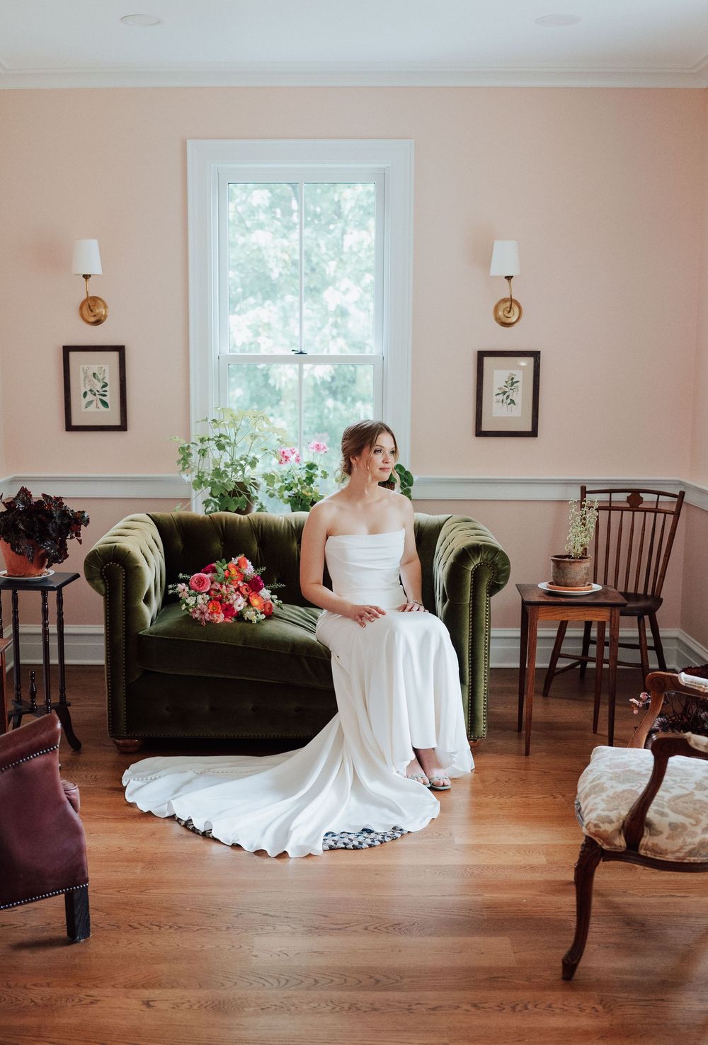 Bride in white gown sits on green velvet sofa holding colorful bouquet in elegant pink room with wood floors.