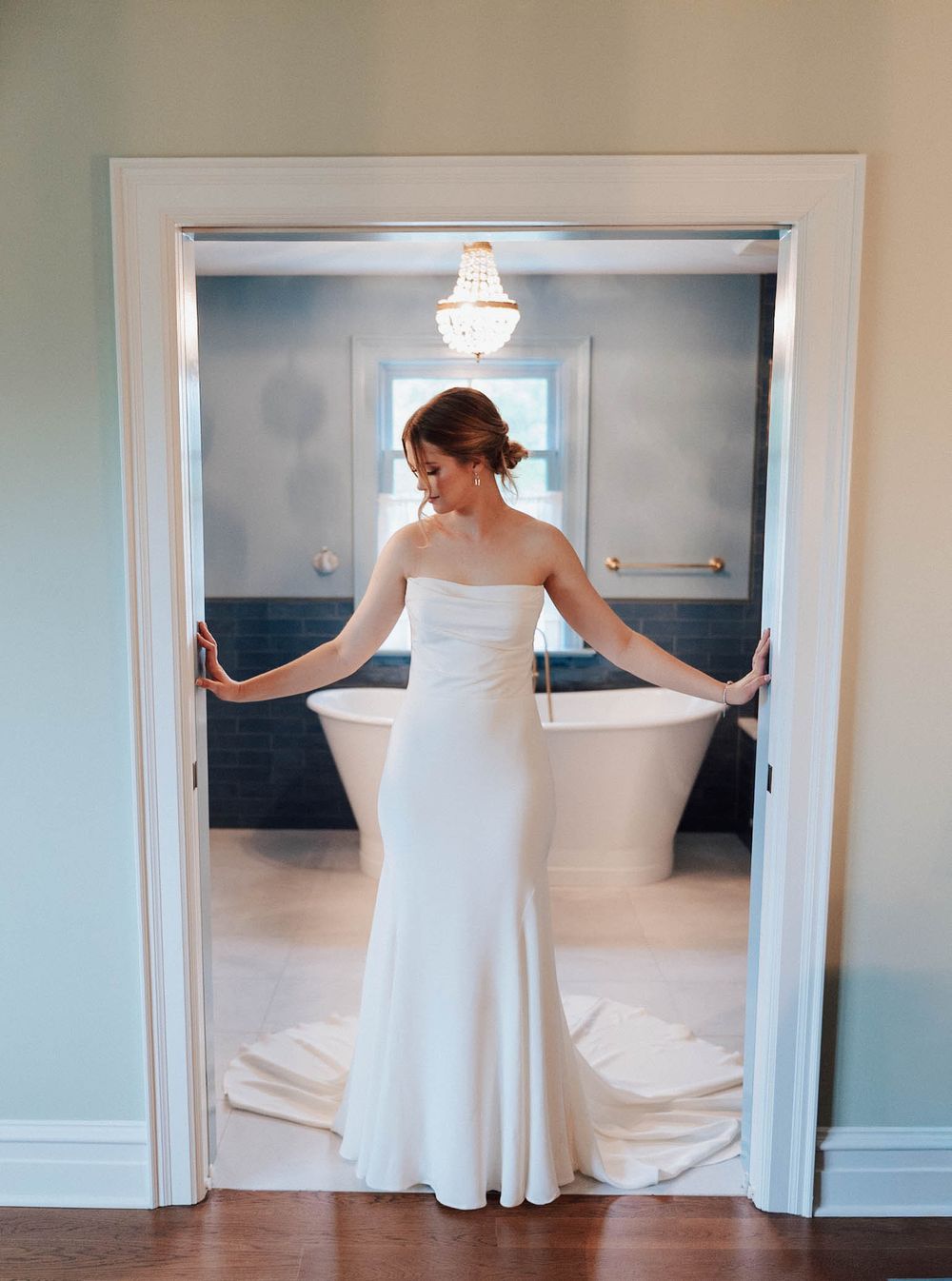 Bride in elegant strapless white gown stands in doorway of luxurious bathroom with freestanding tub and chandelier.