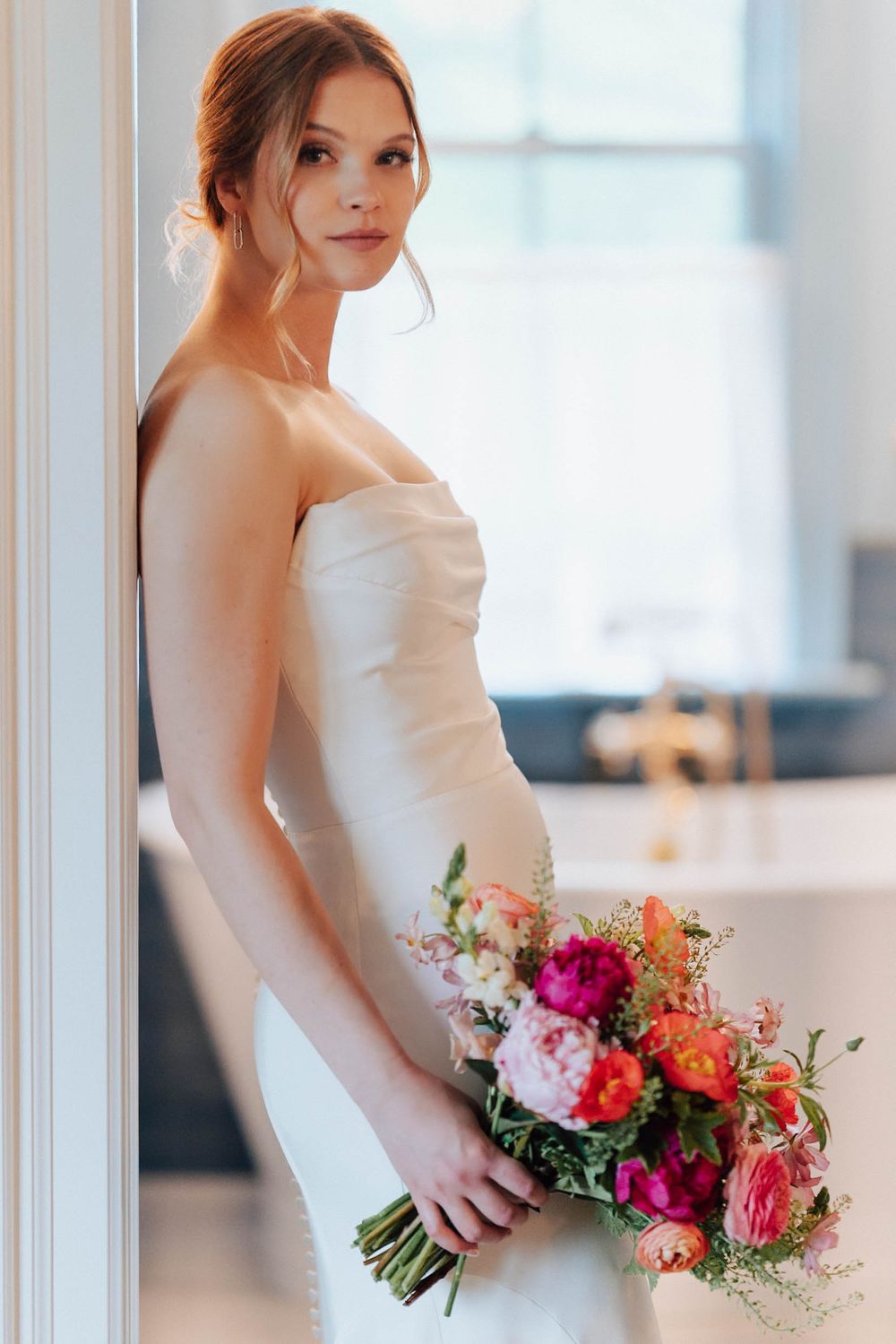 Bride in strapless white gown holding vibrant pink and orange floral bouquet, leaning against white column indoors.