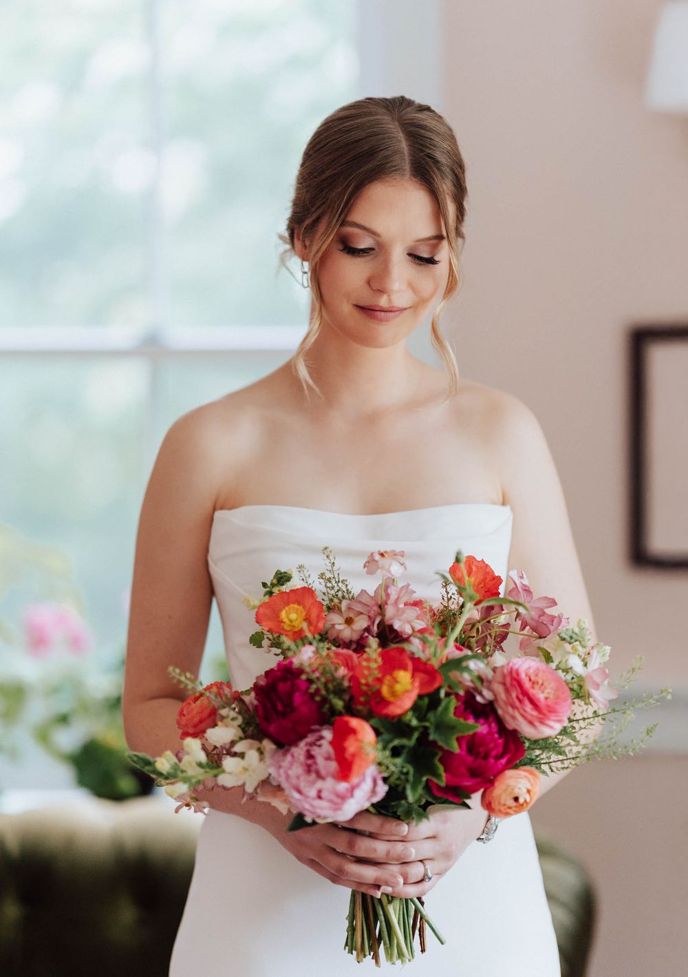 Bride in strapless white gown holding vibrant pink, red, and orange floral bouquet, gazing down near a bright window.