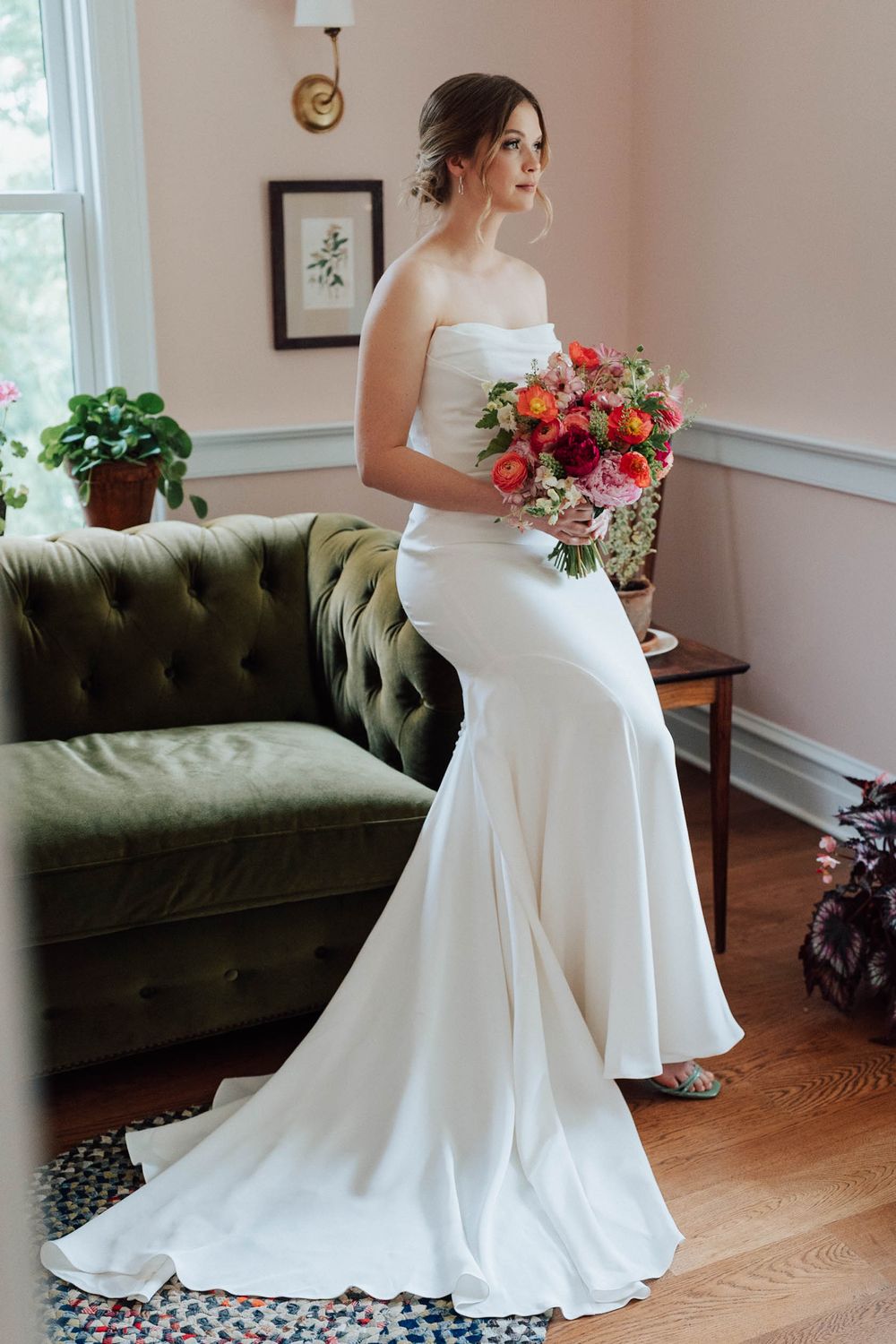 Bride in strapless white mermaid gown holding colorful bouquet stands by green tufted sofa in elegant room.