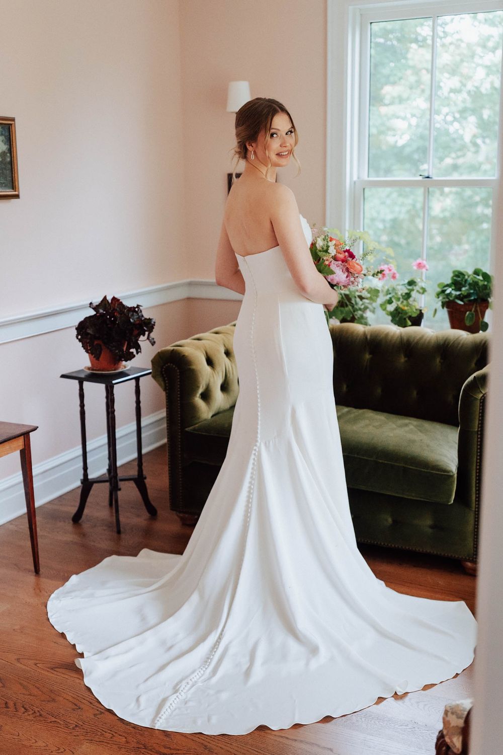 Bride in strapless white wedding gown with train holds bouquet, posing by window near green velvet sofa.