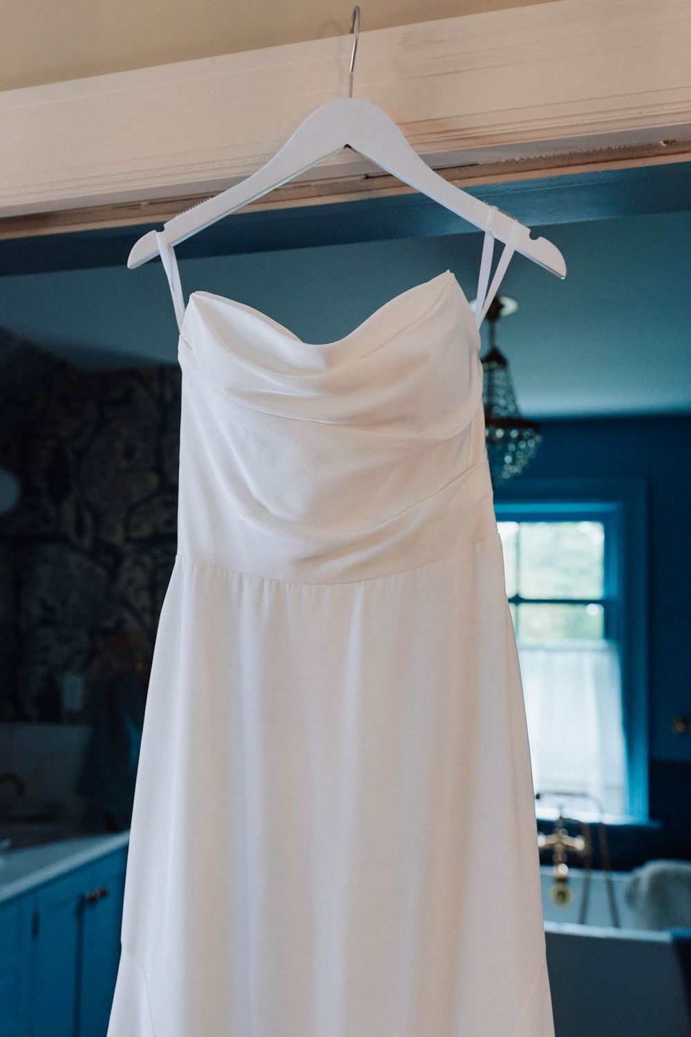 White strapless wedding dress hanging on a white hanger in a blue-walled room with natural light.