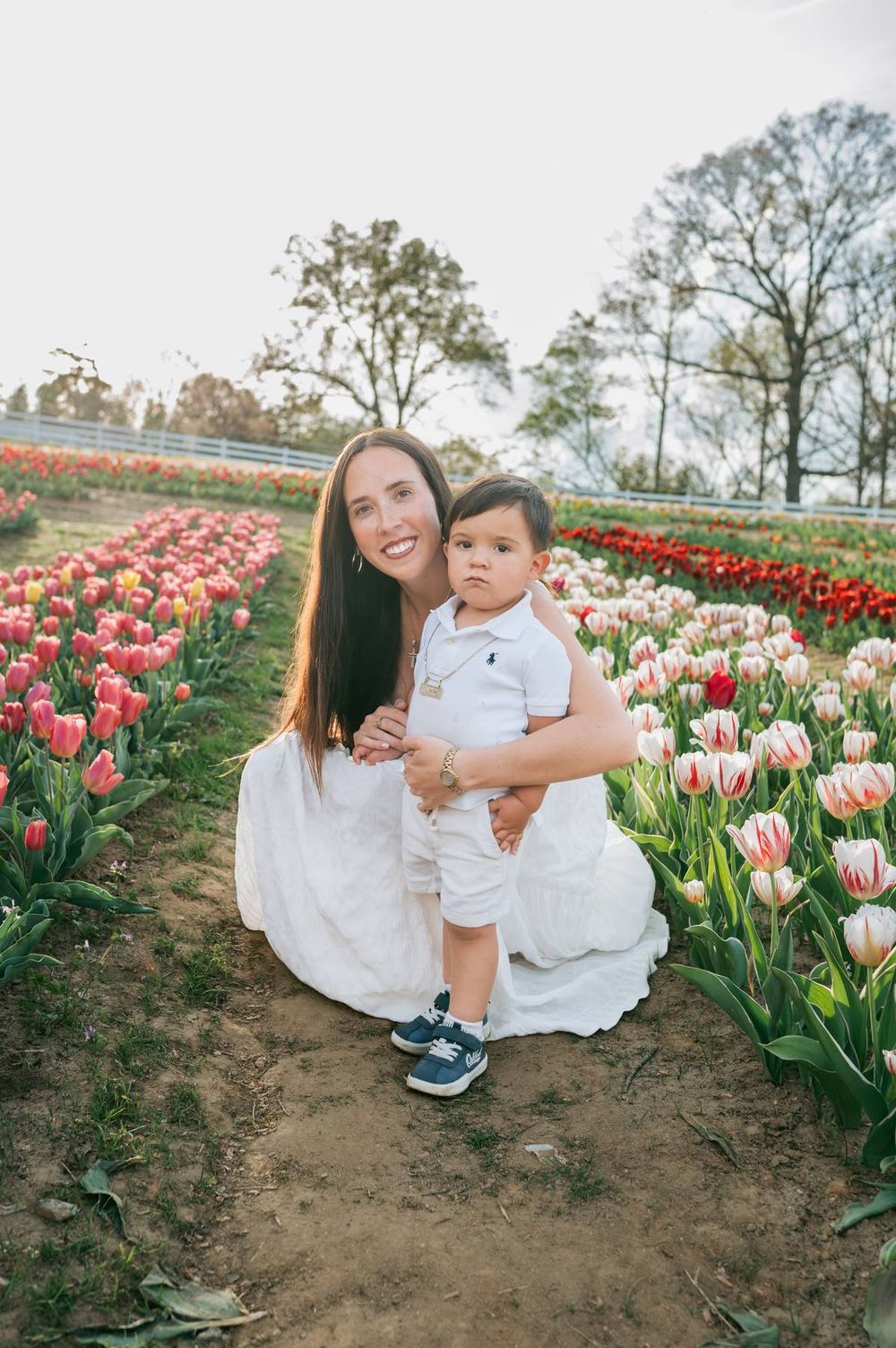 Smiling mother in white dress kneeling with baby boy in tulip field at sunset Asheboro NC