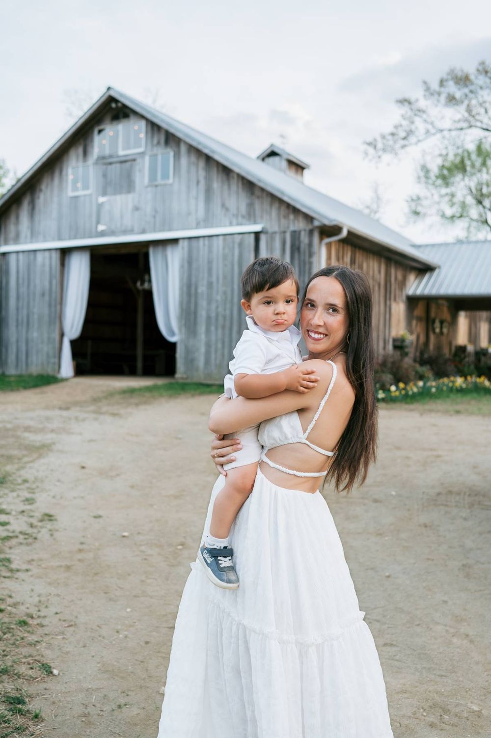 Smiling mother in white dress holding baby boy in front of a rustic wooden barn Lexington NC