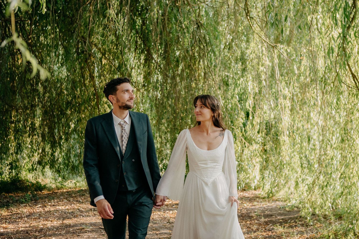 Bride and groom walk hand in hand under a weeping willow tree, she in a white gown, he in a dark suit.