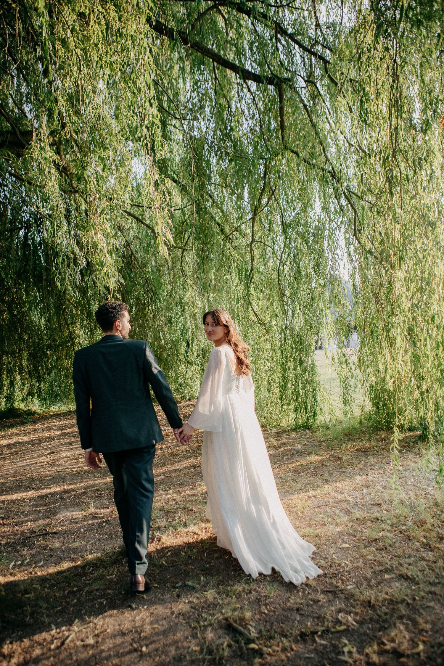 Bride and groom walking hand in hand under a weeping willow tree, bride in flowing white dress.
