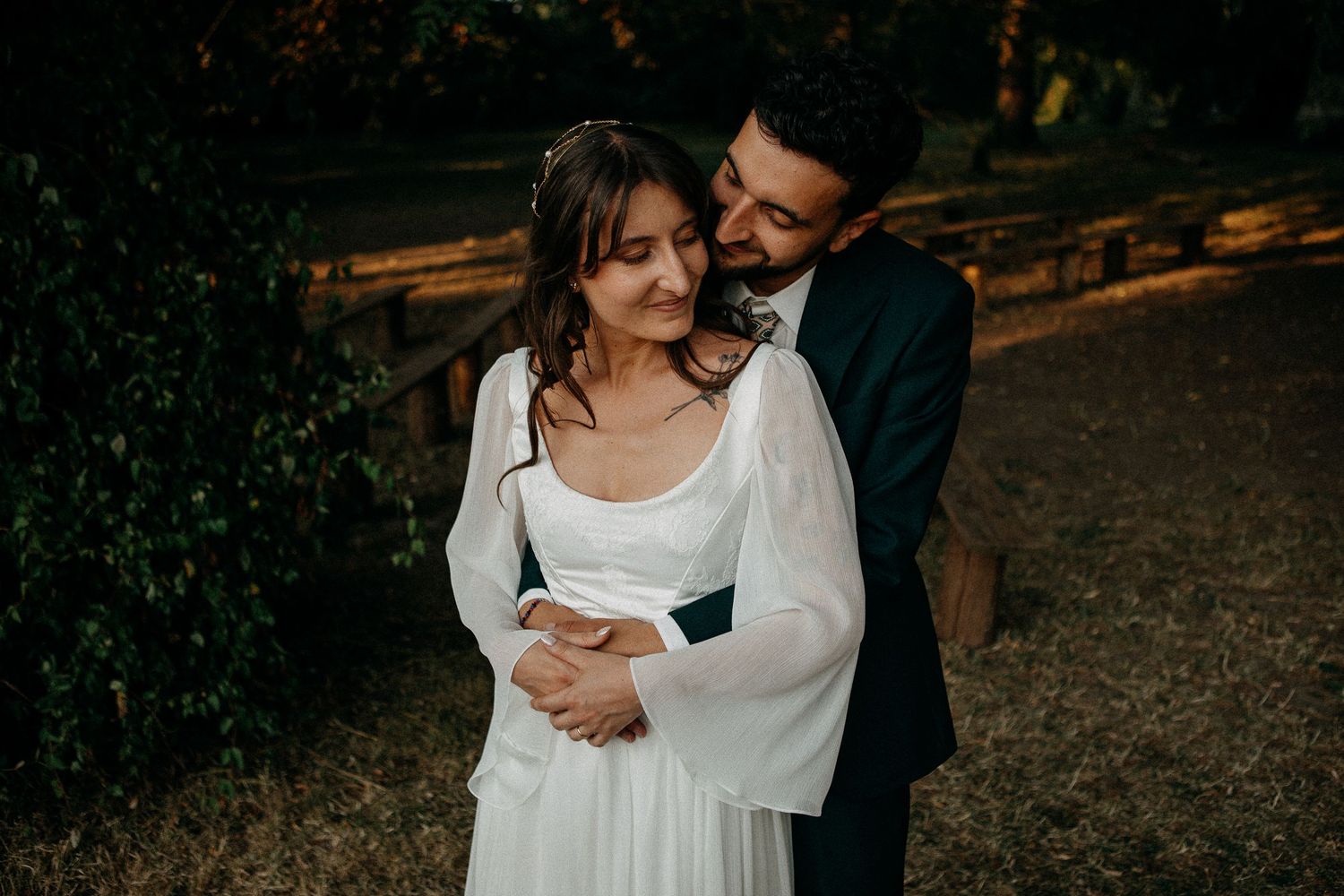 A couple in wedding attire embraces outdoors, the groom in a dark suit holding the bride in a white long-sleeve gown.