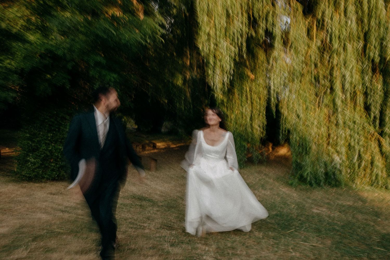 Bride and groom walking under a weeping willow tree in a lush garden, captured with a dreamy motion blur effect.