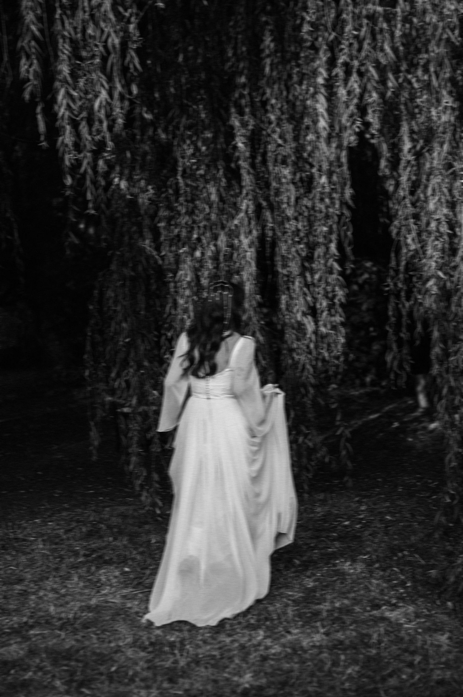 Black and white photo of a woman in a flowing white gown standing beneath a weeping willow tree.