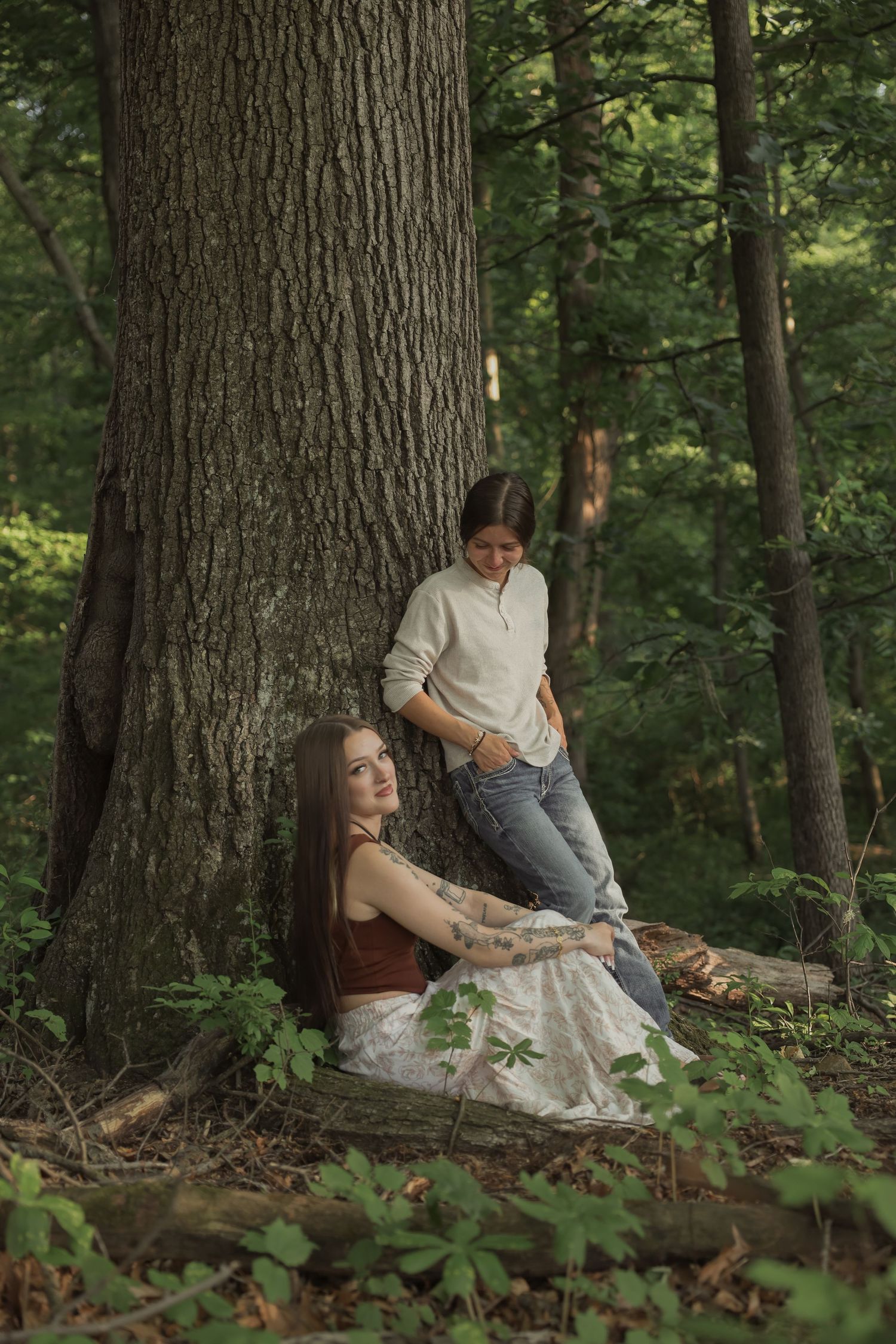 Two women posing by large tree trunk in lush green forest setting.