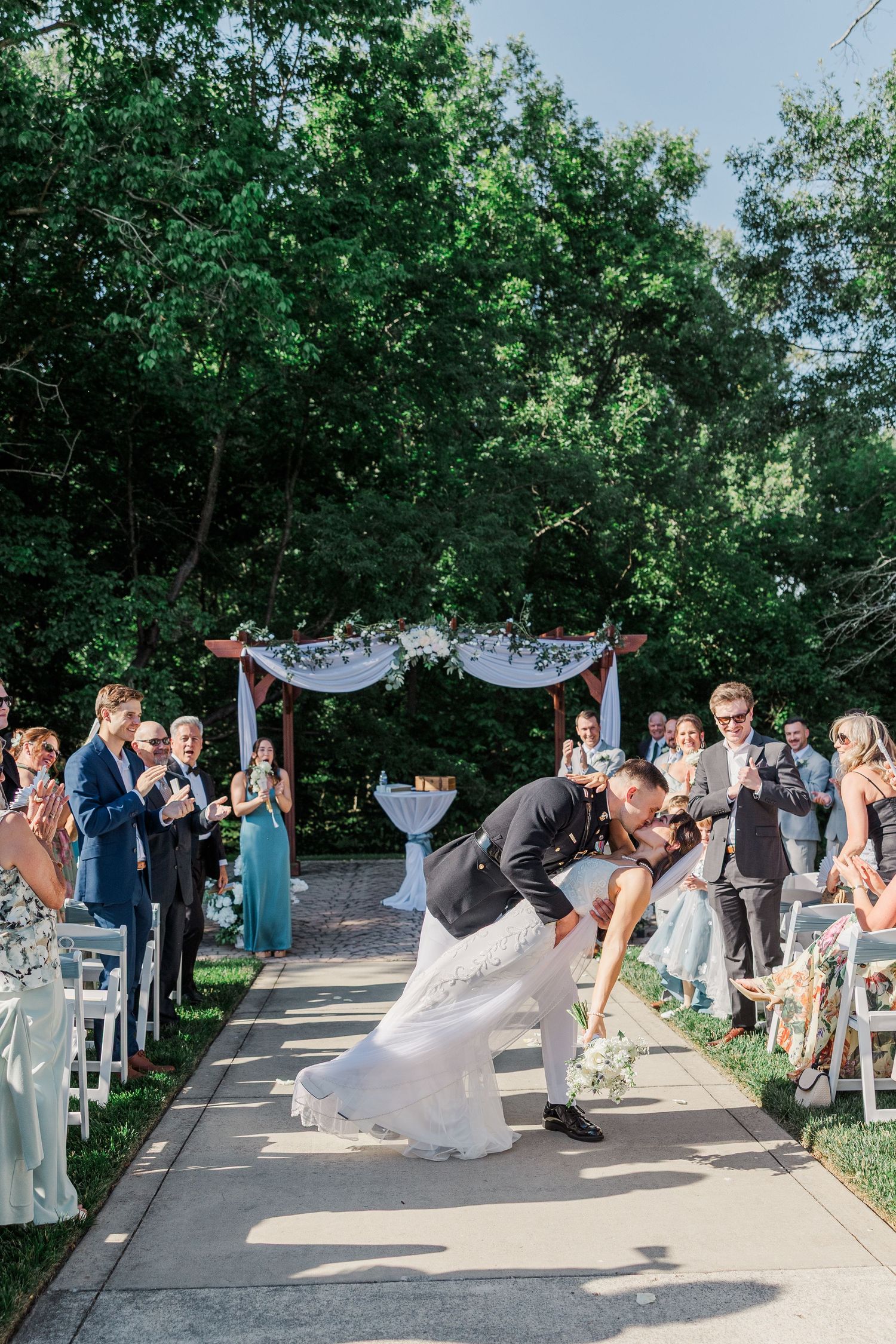 Groom dips bride for a romantic kiss at outdoor wedding ceremony while guests applaud along the aisle.