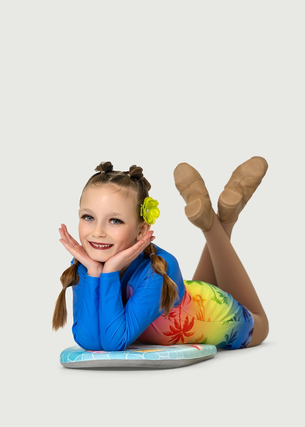 Young girl in blue dance costume with rainbow skirt lying on floor, smiling with hands on cheeks. Dance photo in Regina, Saskatchewan.