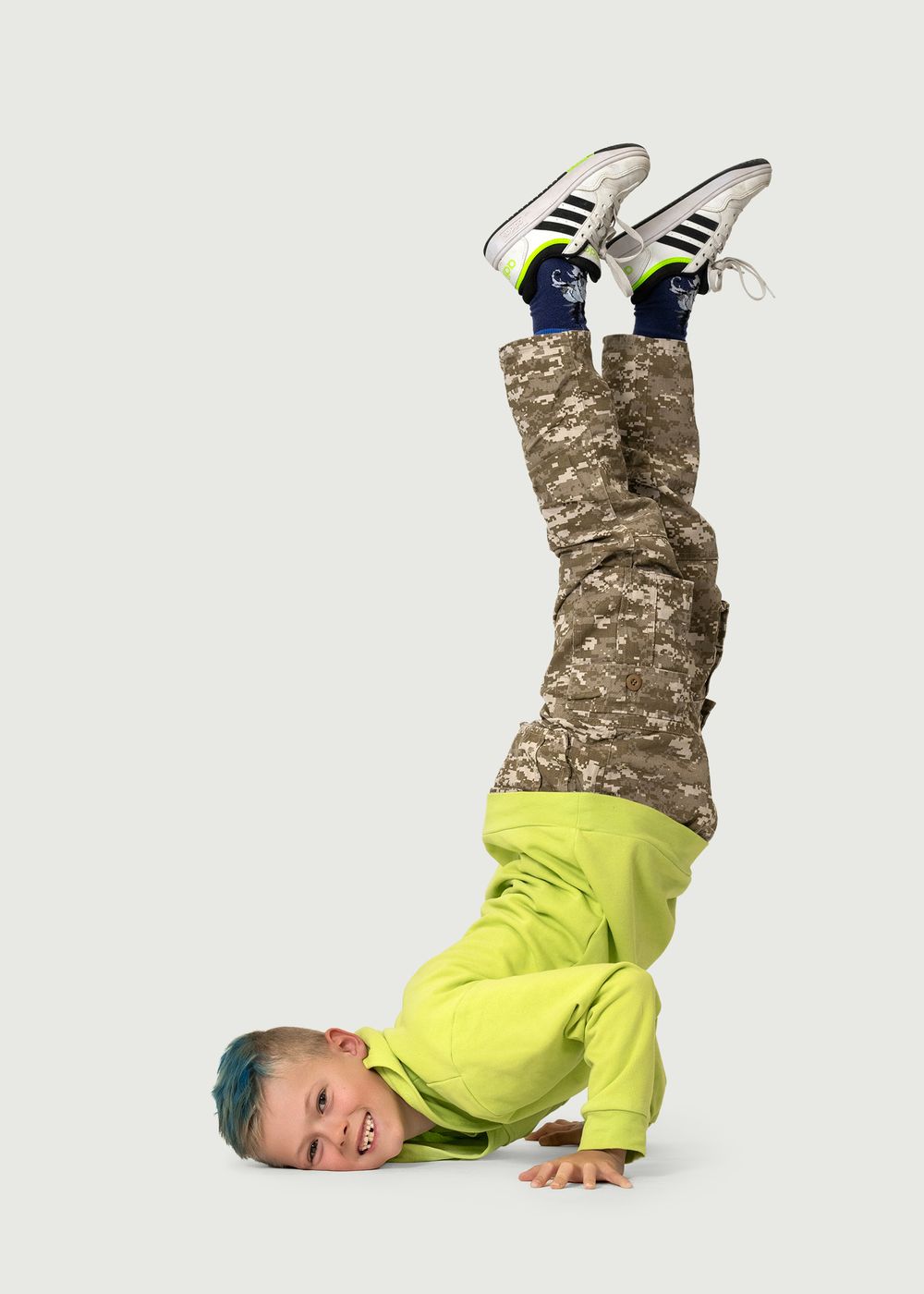 Young boy in neon green hoodie and camo pants doing a headstand, smiling, wearing Adidas sneakers. Dance photo in Regina, Saskatchewan.