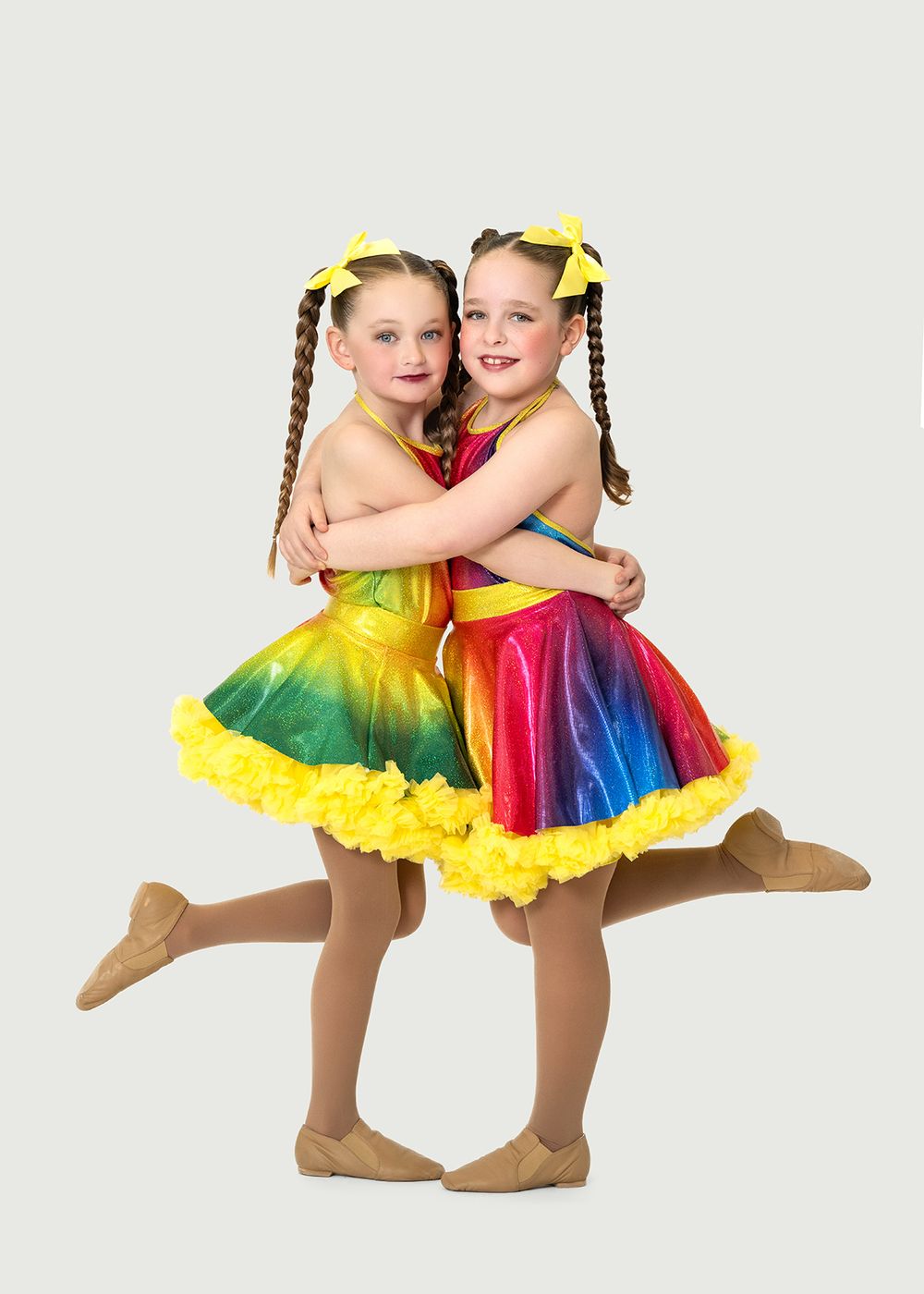 Two young girls in rainbow tutus with yellow bows hugging and posing in a dance recital photo. Dance photo in Regina, Saskatchewan.