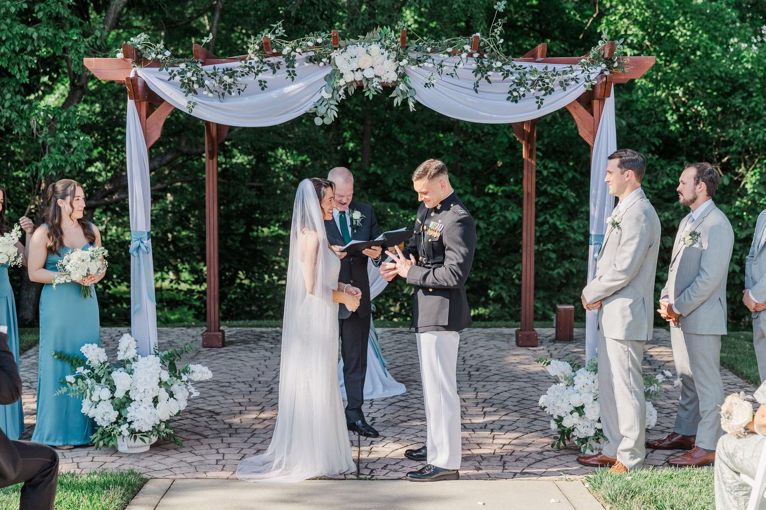 Outdoor military wedding ceremony under floral arch with bride, groom, officiant, and wedding party on brick patio.