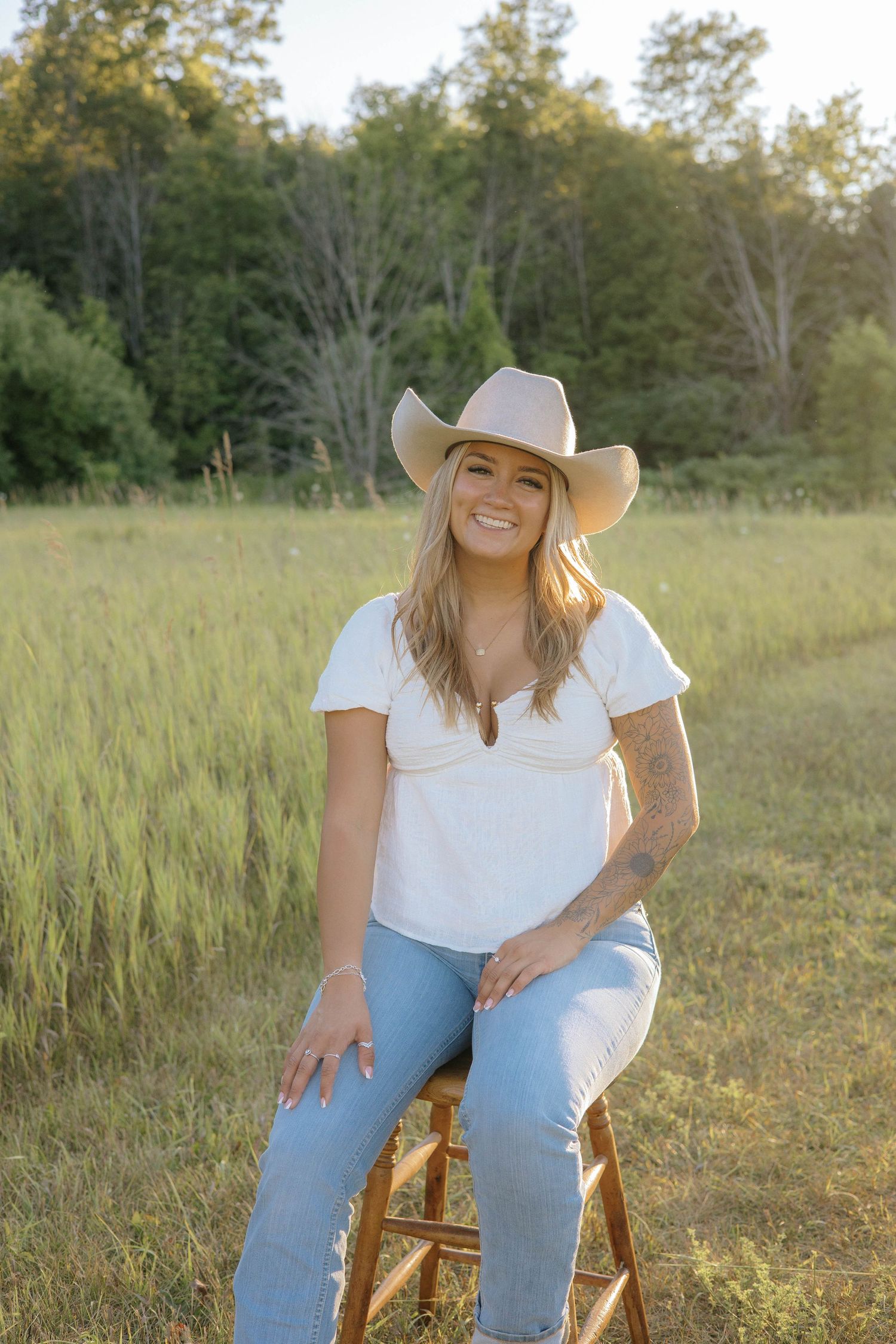 Smiling blonde woman in cowboy hat, white top, and jeans sitting on stool in sunny green field.