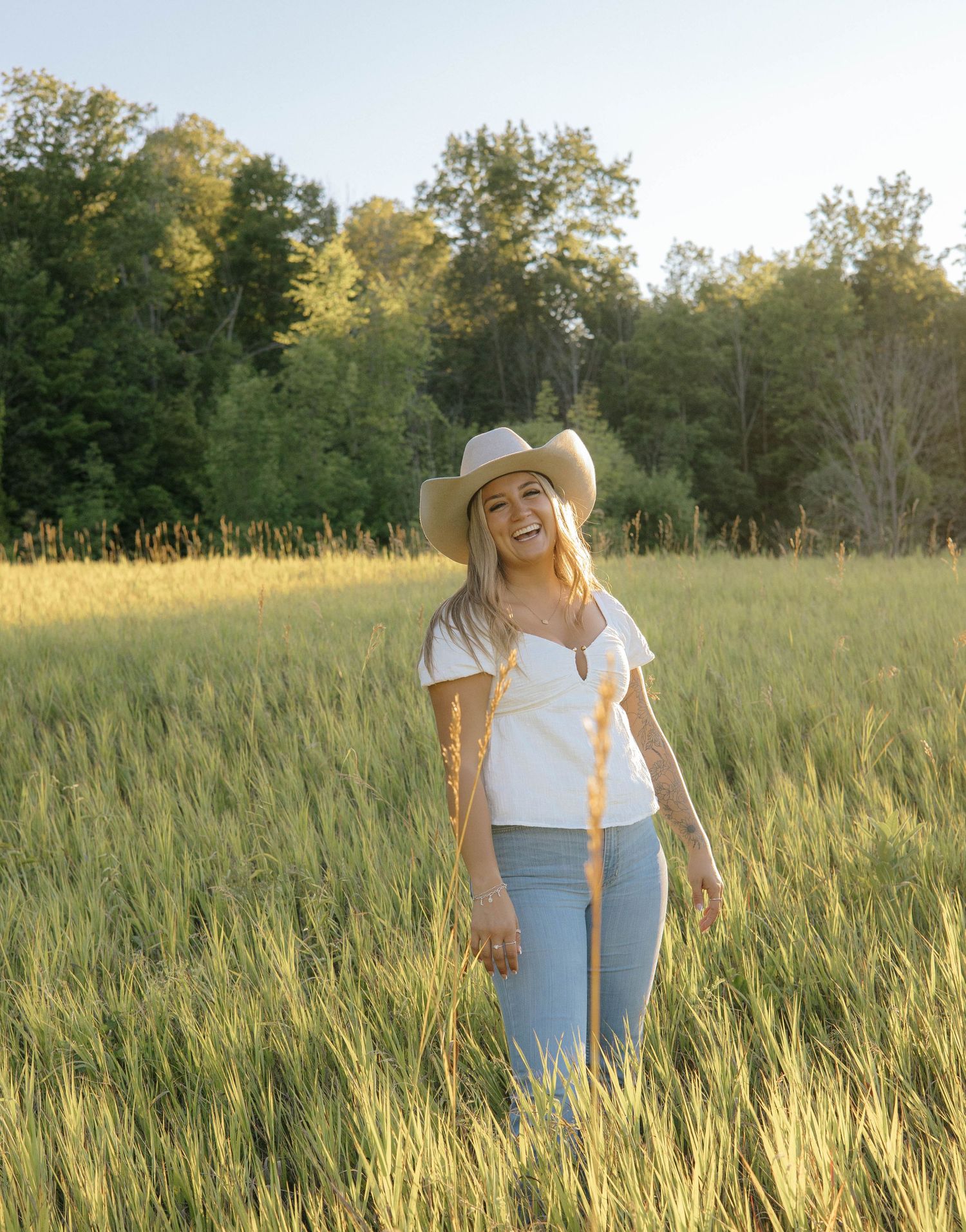 Young woman in cowboy hat and jeans laughing in a sunlit green field with trees in the background.