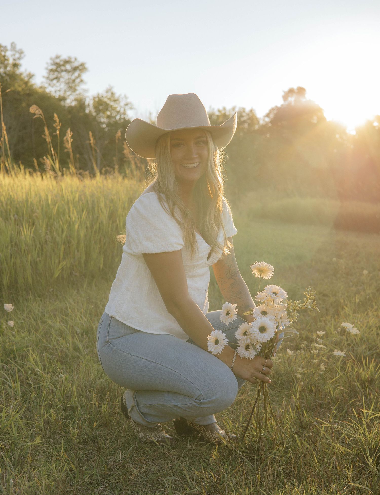 Blonde woman in cowboy hat crouching with white daisies in a sunlit field.