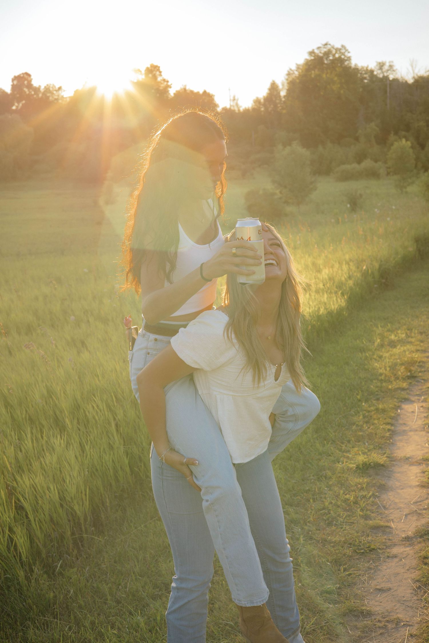 Two women laughing and sharing a piggyback ride in a sunny green field at golden hour sunset.