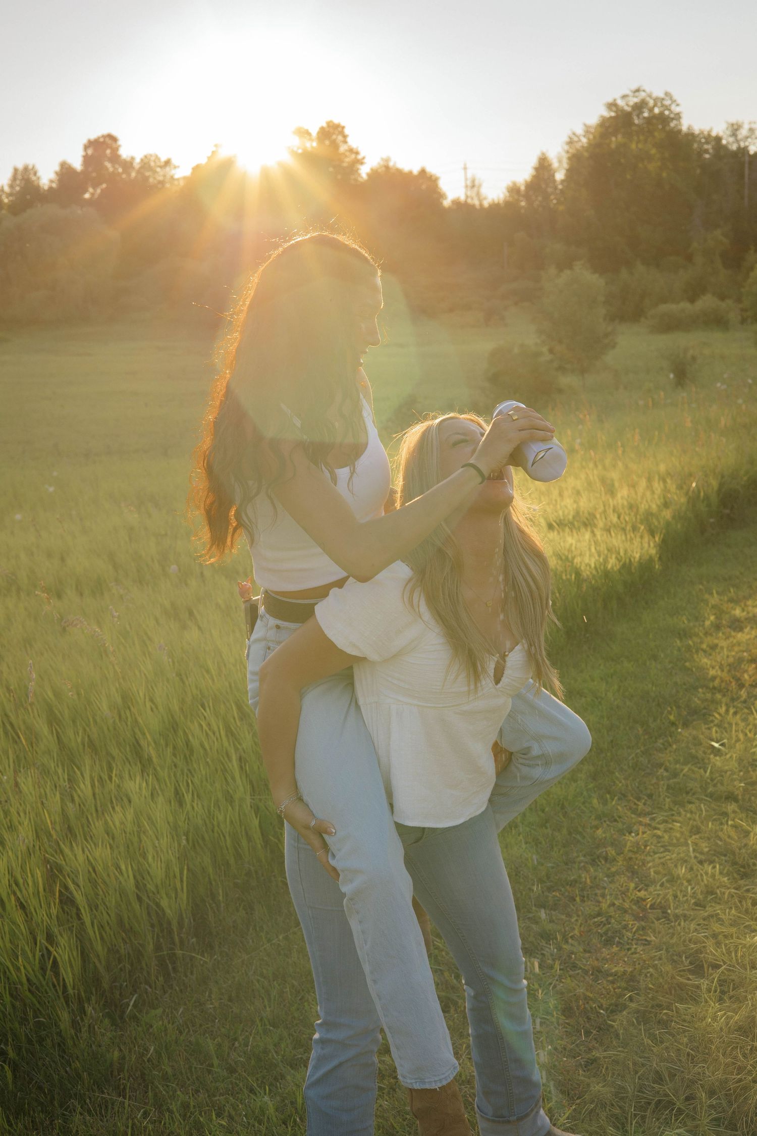 Two women hugging in a golden sunset field with warm sun flare behind them.