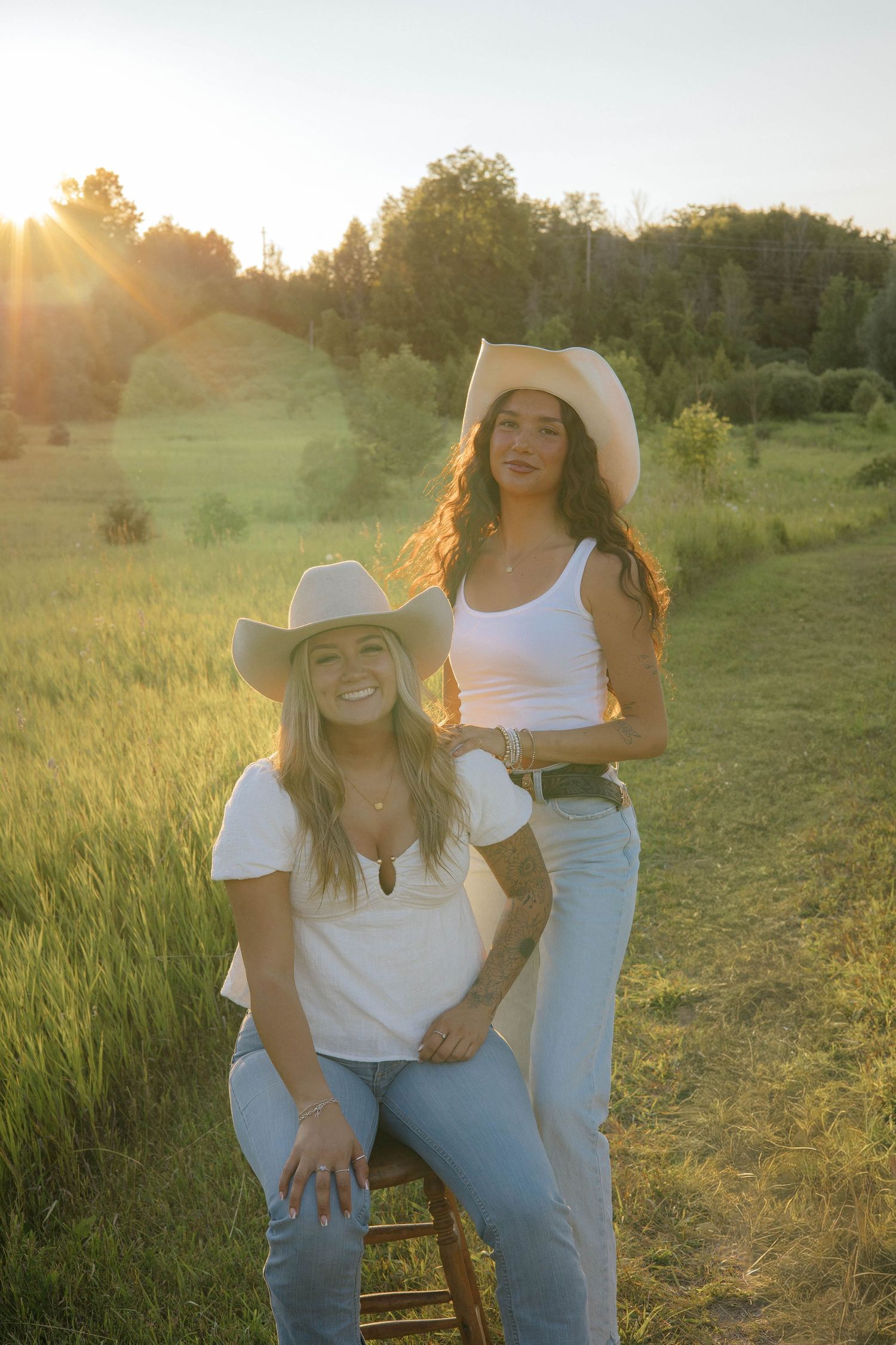 Two young women in white tops, jeans, and cowboy hats pose in a sunlit green field at golden hour.