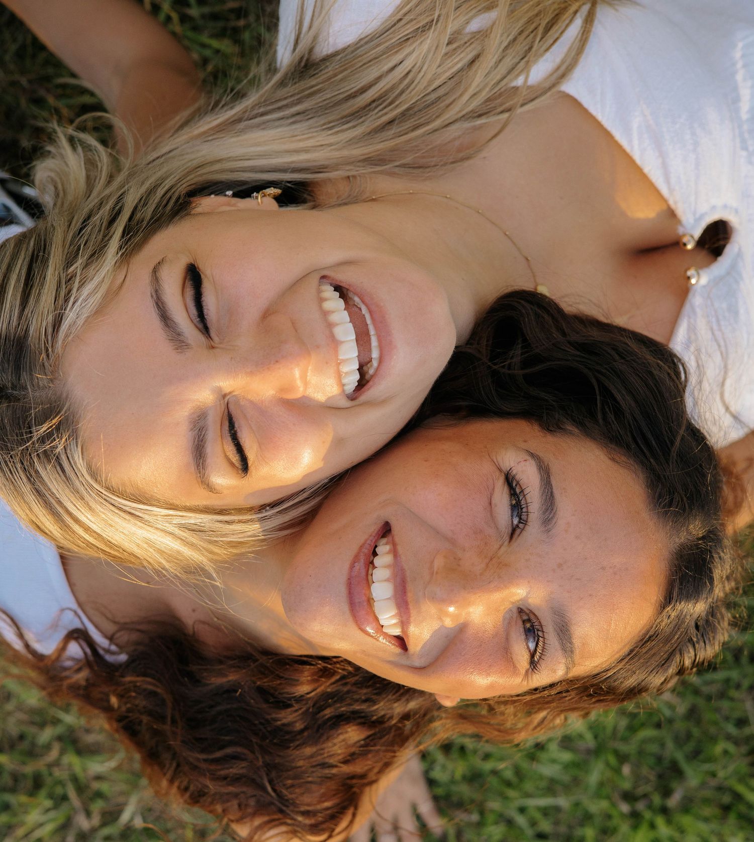 Two smiling women lying on grass, one blonde and one brunette, laughing together outdoors on a sunny day.