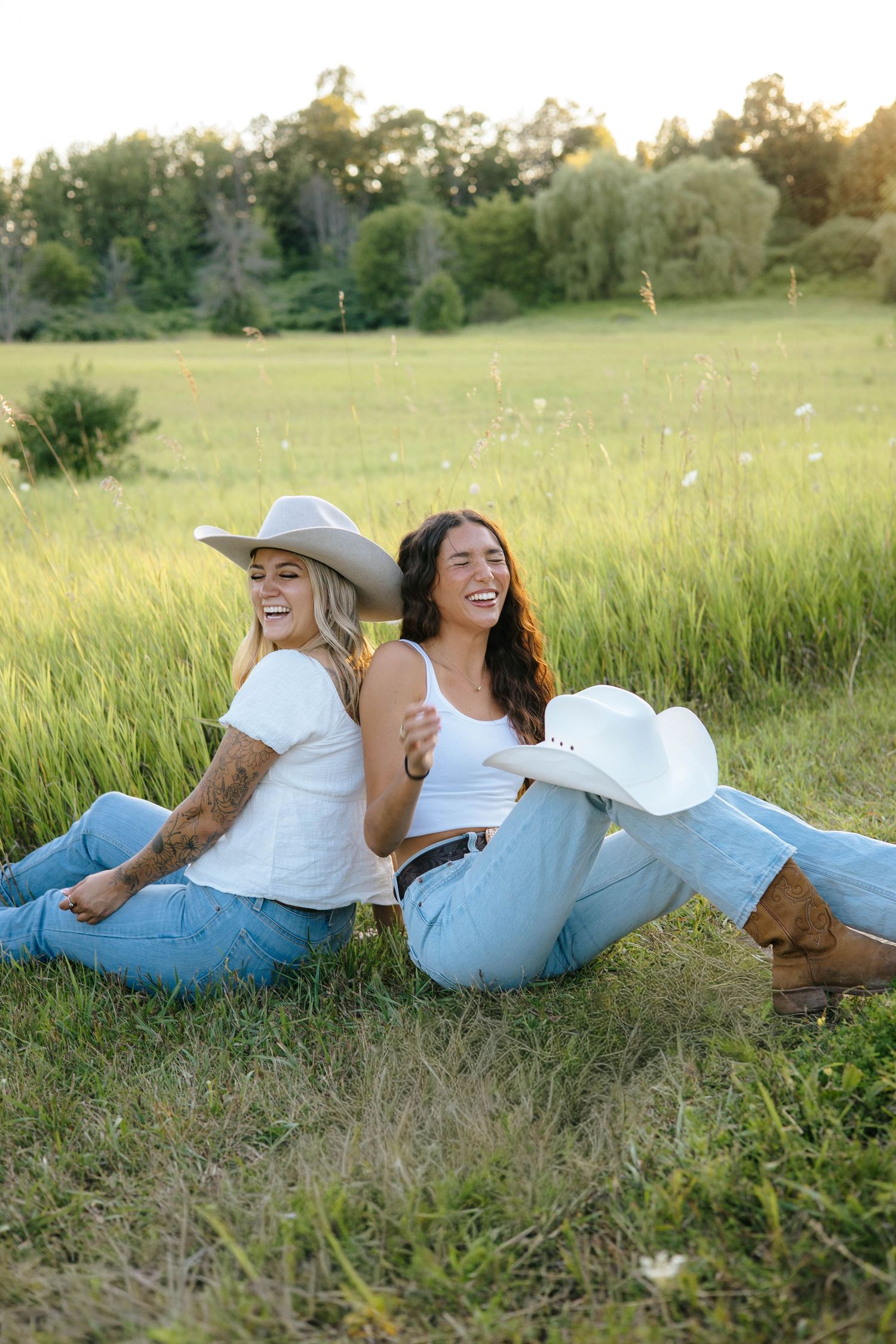 Two smiling women in white tops sitting back-to-back in a green grass field.